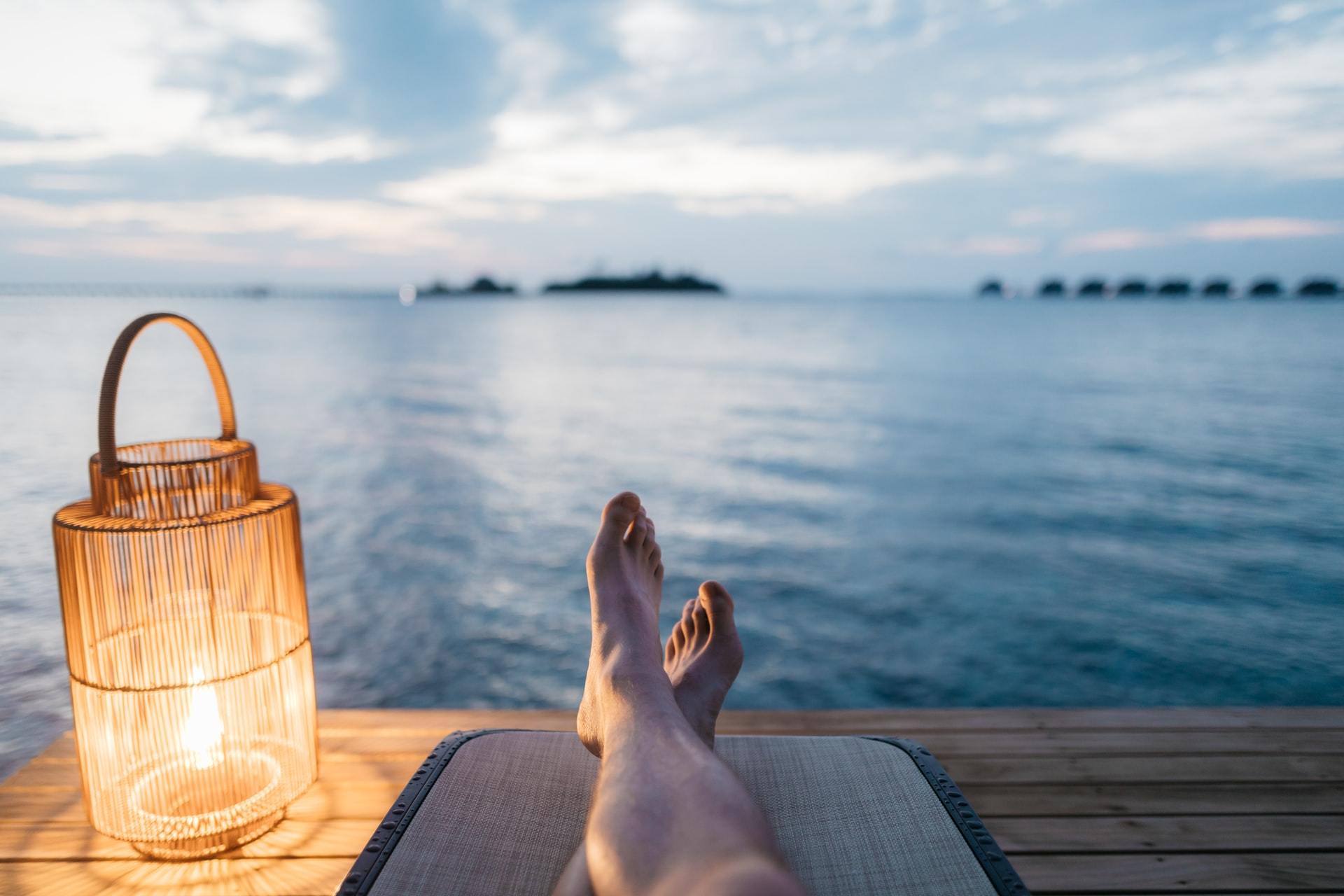 Lit lantern candle beside relaxing feet by calm water at dusk