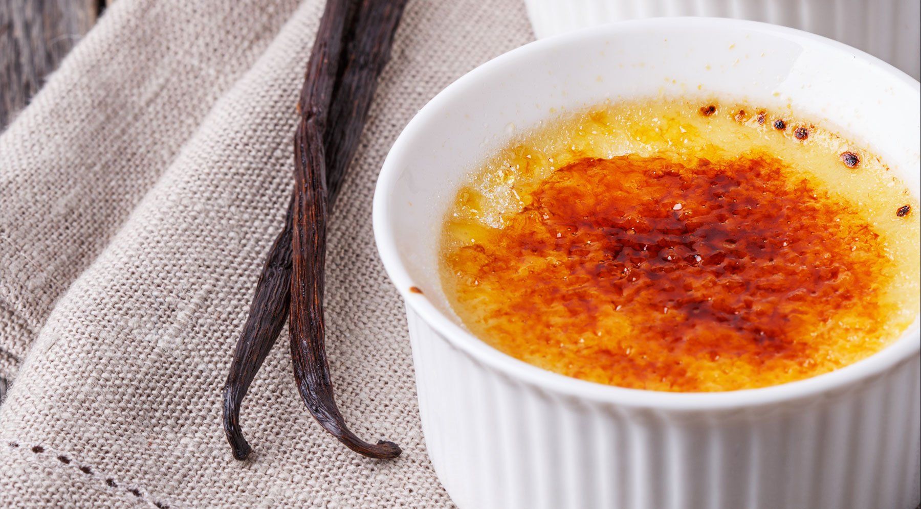 Crème brûlée ramekin with caramelized top and vanilla pods on linen