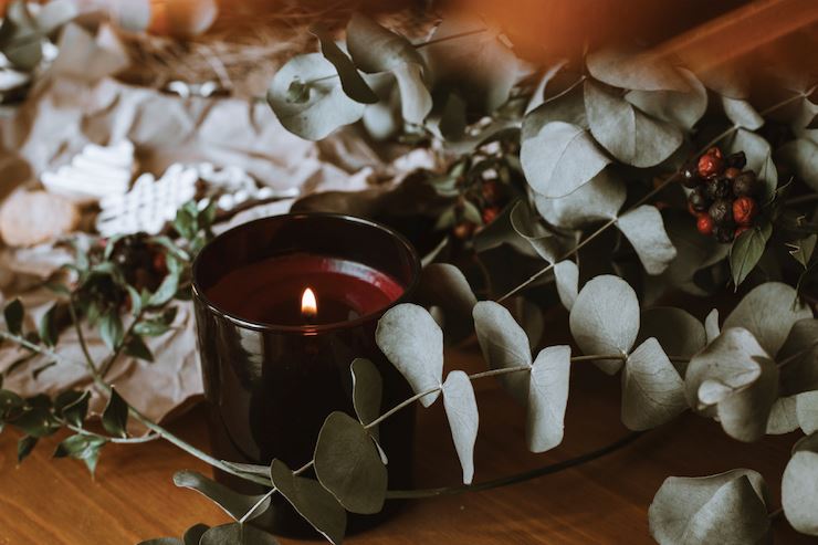 Lit candle surrounded by eucalyptus leaves and dried botanicals on wood