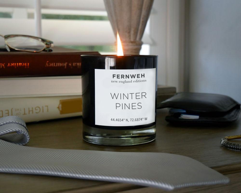 Lit Fernweh Winter Pines candle on a desk with books and glasses